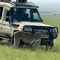 Lioness stands in tall grass beside a safari vehicle, with a vast green plain and distant hills behind her under a bright sky.